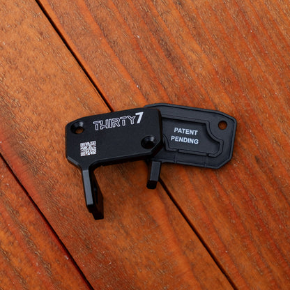 A black metal lock pick tool with 'THIRTY7' and 'PATENT PENDING' printed on it, resting on a brown wooden plank surface.