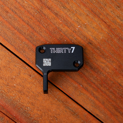 A black metal accessory engraved with the brand name 'THIRTY7' and a QR code, resting on a warm brown wooden surface.
